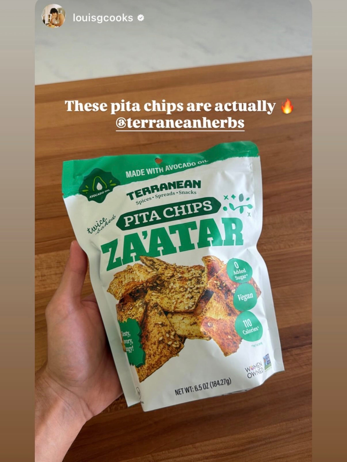A hand holds a bag of Terranean Herb Co. Zaatar Pita Chips on a wooden cutting board, with a social media post overlay at the top.