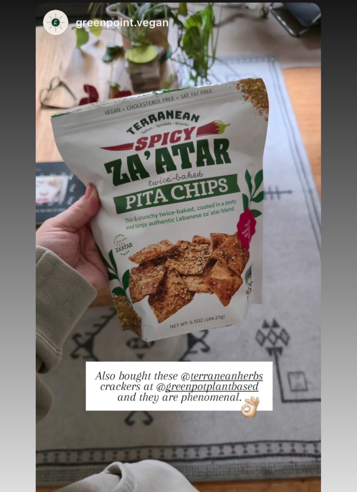 A hand holds a bag of Terranean Spicy Za'atar flavored pita chips in a home setting. The bag is white with green and red text and a photo of pita chips on the front.
